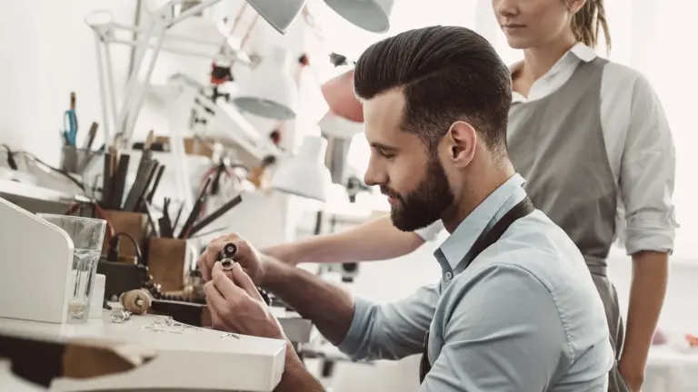 Ein Mann arbeitet an einem Schmuckstück in einer Werkstatt, während eine Frau im Hintergrund assistiert.