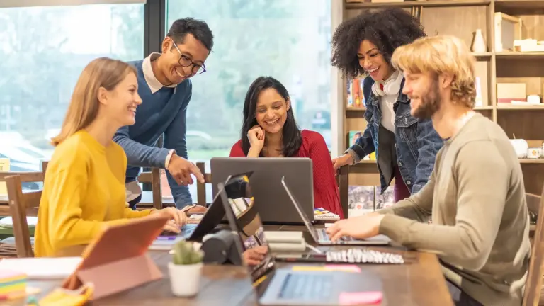 Sechs Personen sitzen an einem Tisch und arbeiten gemeinsam an Laptops und Tablets.