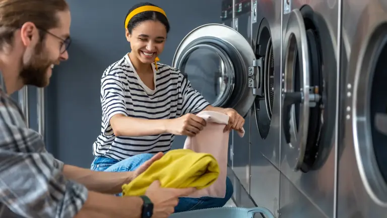 Zwei Personen in einer Wäscherei, eine Person hält einen gelben Pullover, die andere zieht Kleidung aus einer Waschmaschine.