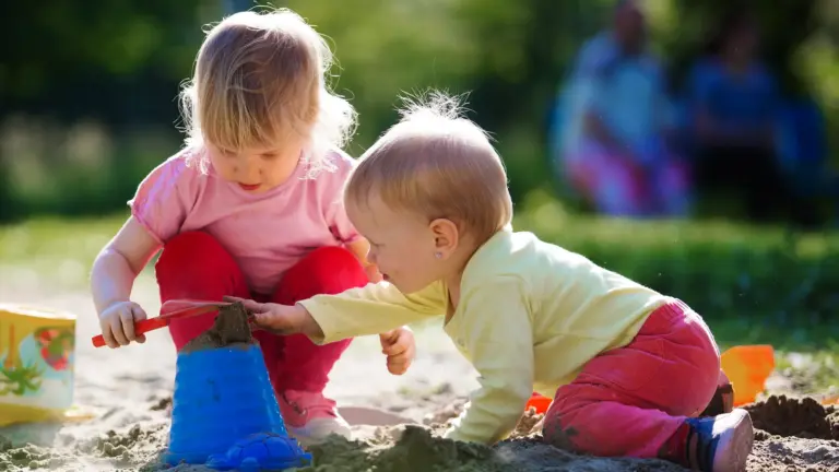 Zwei kleine Kinder spielen im Sandkasten, eines mit einer Schaufel, das andere mit einem Sandturm.